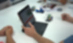 People collaborating at a white table, focusing on a tablet displaying LEGO® designs. Small LEGO® models and pieces are scattered around the workspace, depicting an active design or brainstorm session.