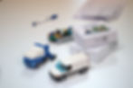 A close-up of various LEGO® vehicle models on a white table, including a blue truck and a white van, accompanied by product boxes labeled 'SCAN TO BUILD.' A pen and blurred documents are also visible in the background.